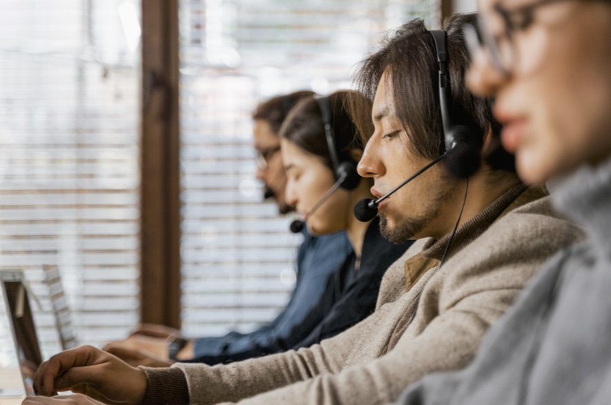 Vier Menschen mit Headset sitzen in einer Reihe an Bildschirmen und telefonieren.