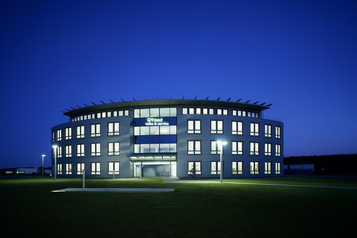Das Firmengebäude von QVmed sales & service, abends beleuchtet, mit dem Schriftzug QVmed sales & service an der Glasfront